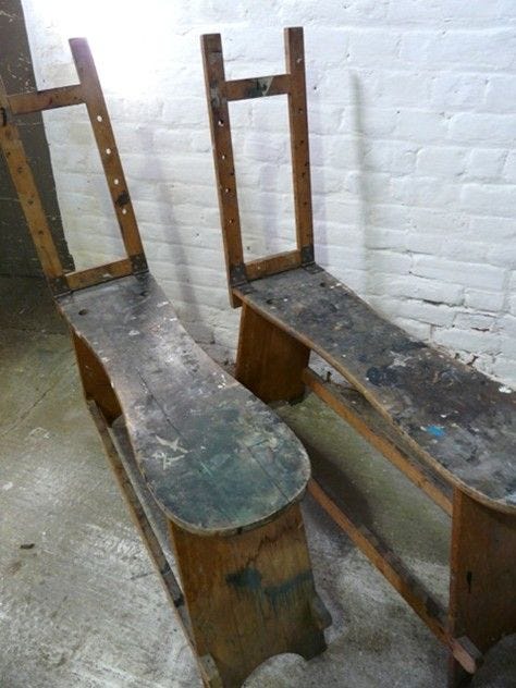 Two unfinished wooden chairs with a rustic design, positioned side by side, in a dimly lit room with a white brick wall.