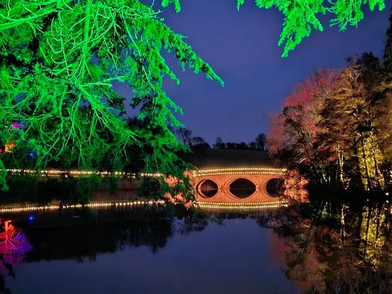 lit up arched bridge over the lake reflected through a frame of lit up tree branches