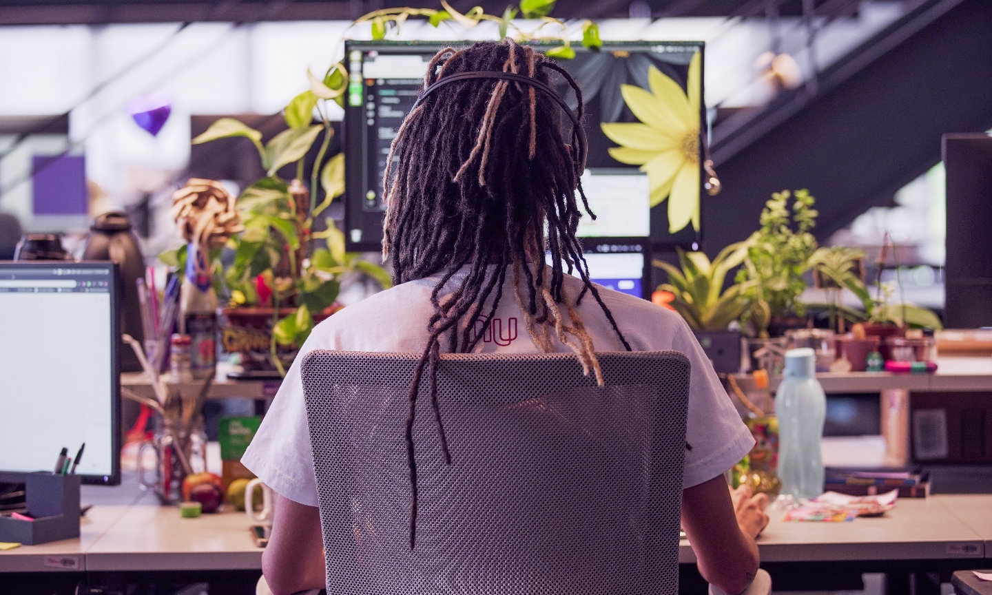 A young man with dreadlocks seen from behind works in his computer, that is surrounded by plants.