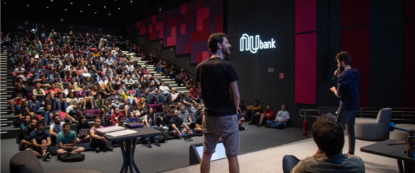 Two young men speak on stage in front of the Nubank's auditorium
