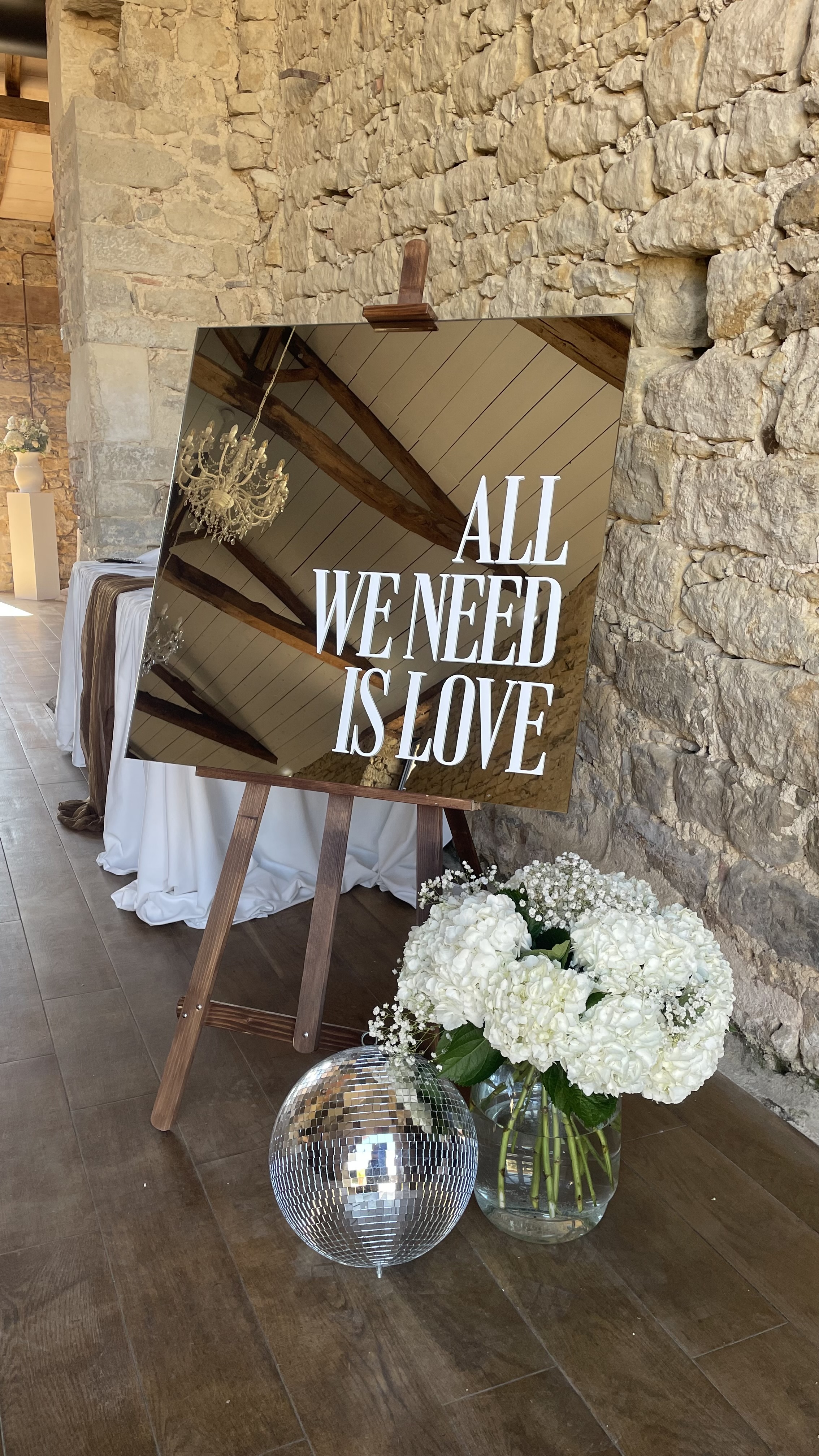 Panneau « All we need is love » sur chevalet en bois, décoration de mariage devant un mur en pierre, organisé par Bulle Wedding Planner