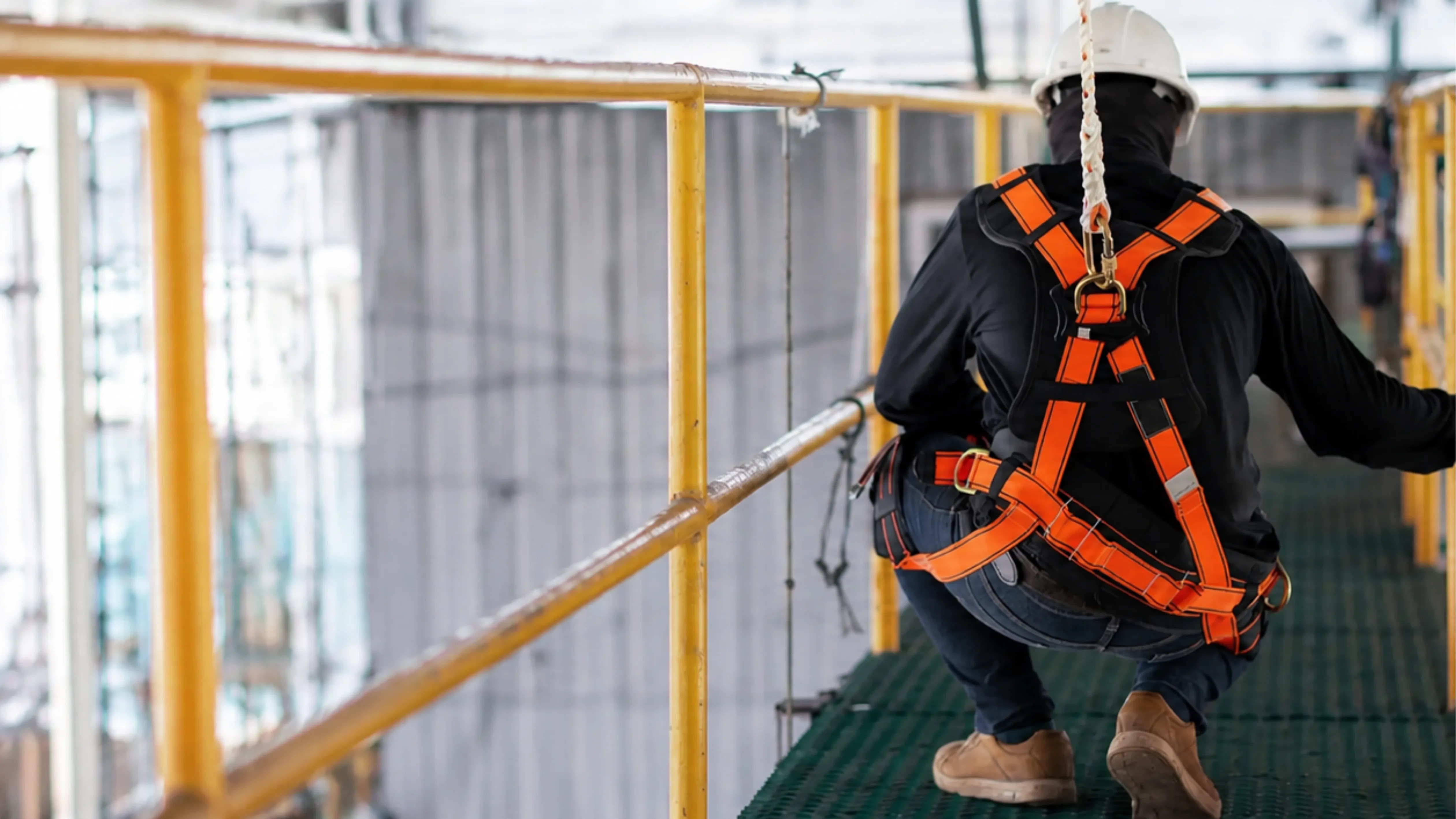 Photorealistic wide image of a worker crouching on an elevated green grated industrial walkway, seen from behind while wearing a white hard hat, dark clothing, tan work boots, and a bright orange fall-protection harness attached to an overhead safety lanyard. Yellow safety railings line the catwalk inside a softly lit industrial facility.