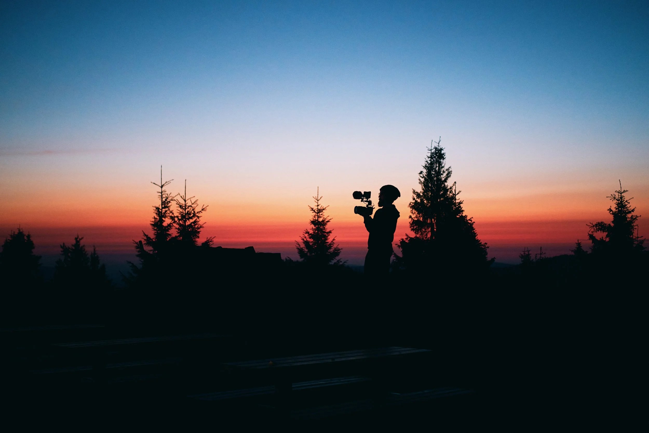 Osoba z kamerą na tle kolorowego zachodu słońca i drzew, scena outdoorowa, filmowanie na zewnątrz.