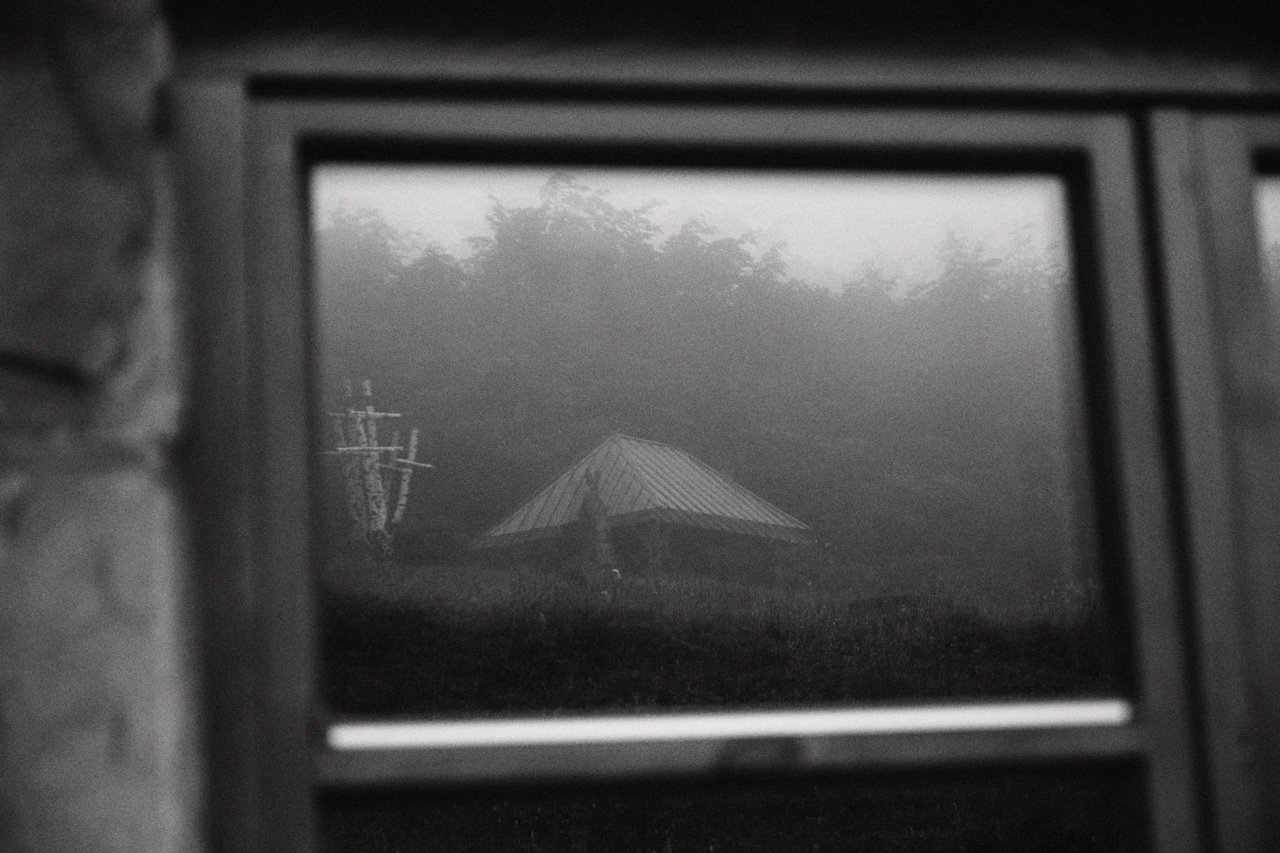 Widok na drewniany dom przez okno, otoczony mgłą i naturą, w stylu czarno-białej fotografii artystyc.