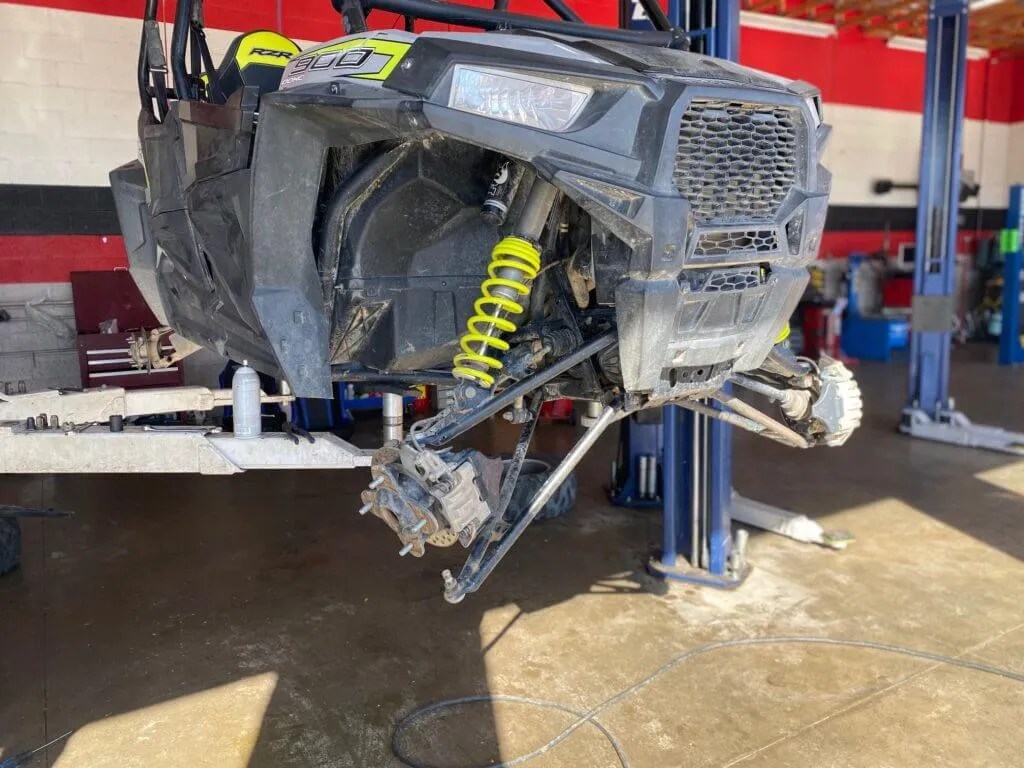 An off-road vehicle is lifted on a mechanic's platform, showcasing the front suspension and yellow shock absorber.