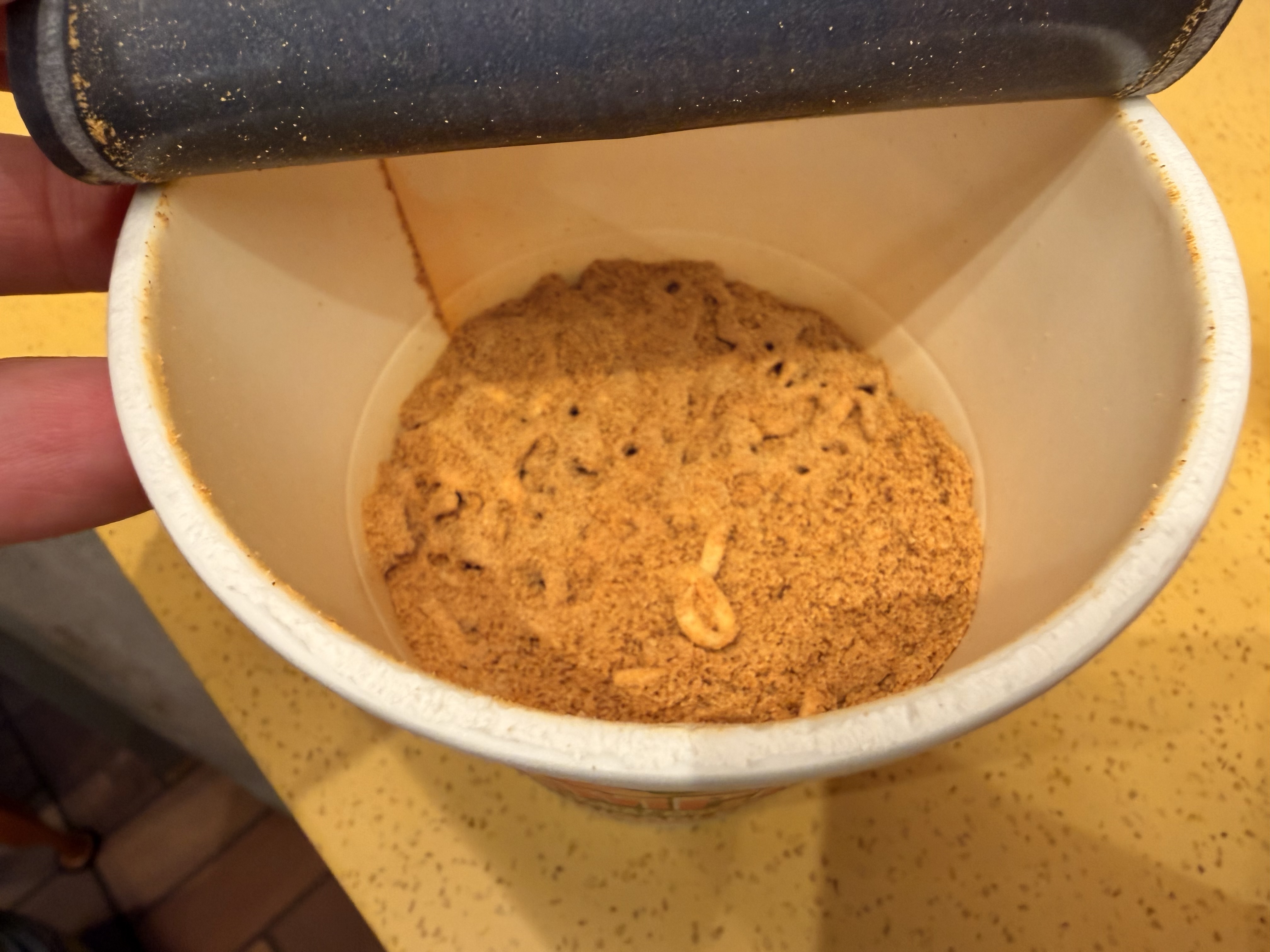 An open container holding a small amount of brown powder, possibly seasoning or spice, with a person's hand visible near the edge.