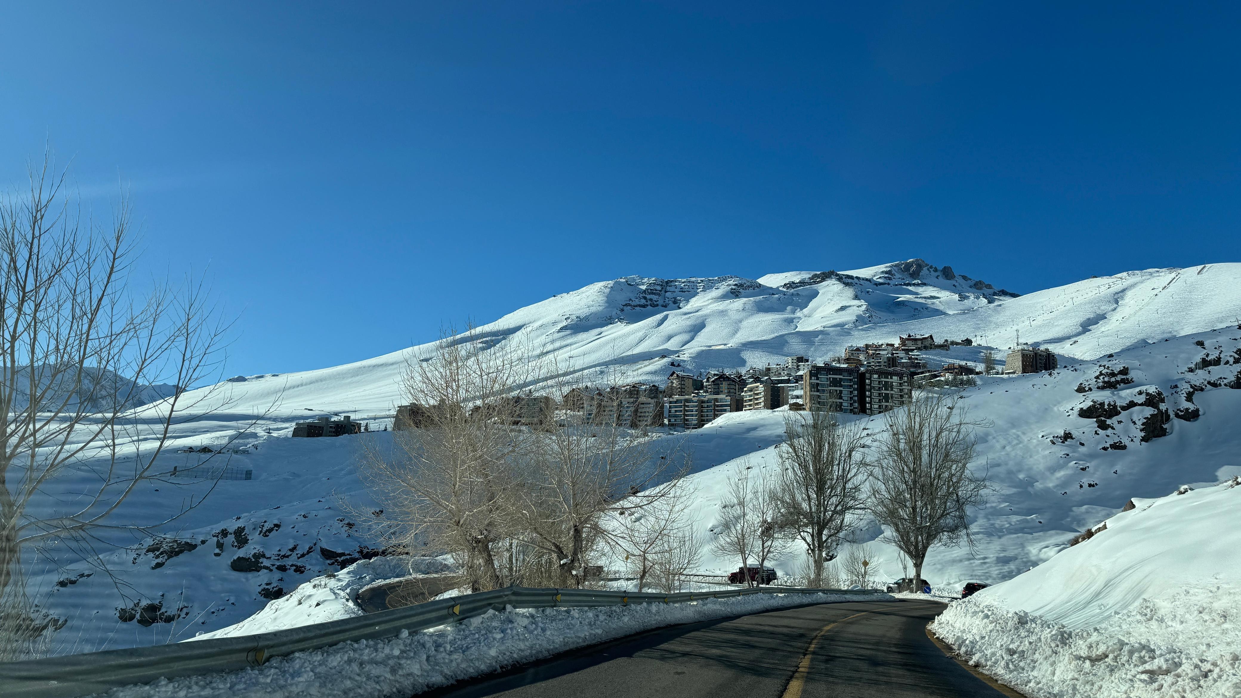 A imagem mostra a estrada com vista para o vilarejo de La Parva, no Chile, em um dia de sol e com o entorno todo coberto de neve.