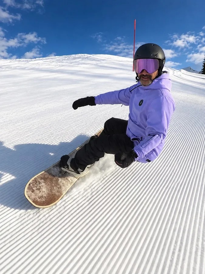 Victor Ramos fazendo uma curva de snowboard em pista de neve recém-preparada, sob céu azul