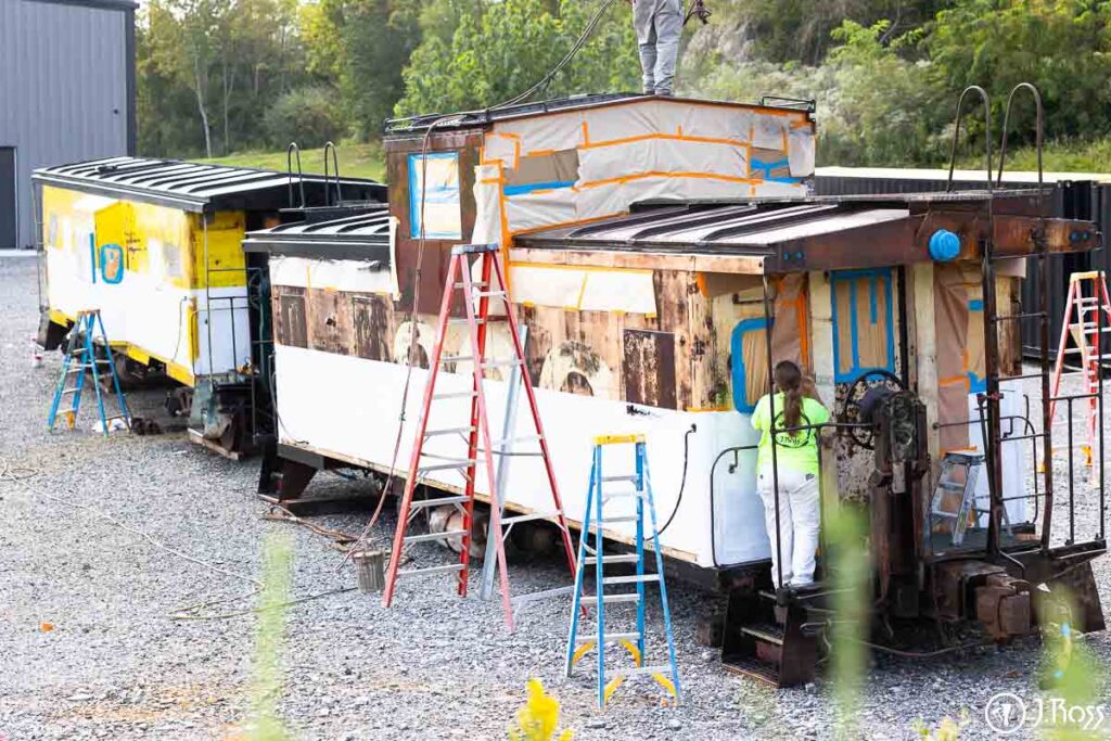 Two cabooses side by side at J. Ross headquarters with crews applying white stripe and black roof coatings during restoration