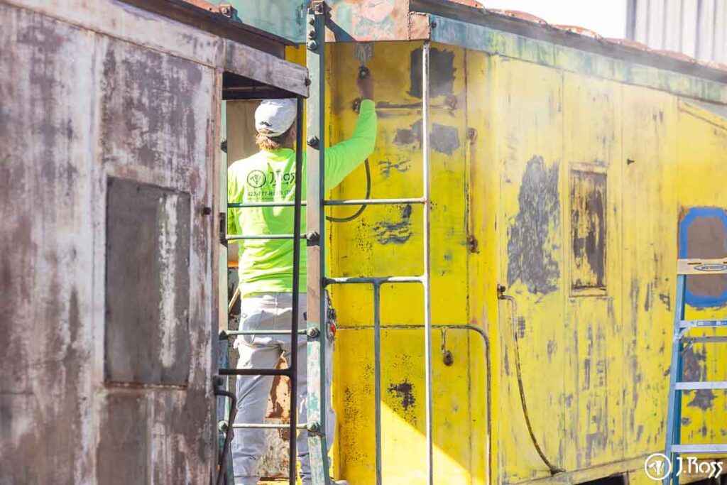 J. Ross Painting crew applying Sherwin-Williams Macropoxy 920 primer to yellow caboose exterior during restoration