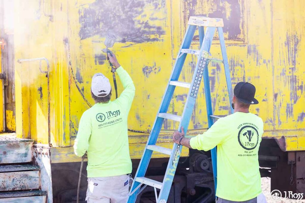 Painters spraying sherwin-williams macropoxy 920 on caboose exterior at J. Ross Painting & Drywall shop