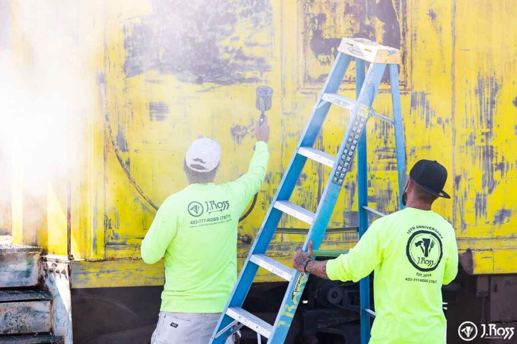 J. Ross Painting team spraying industrial coatings on vintage yellow caboose exterior during restoration process