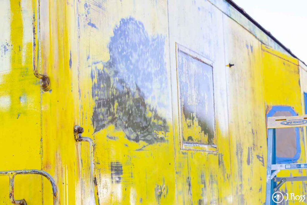 Detail of yellow caboose panel with sanding and grinding marks revealing steel beneath old paint layers