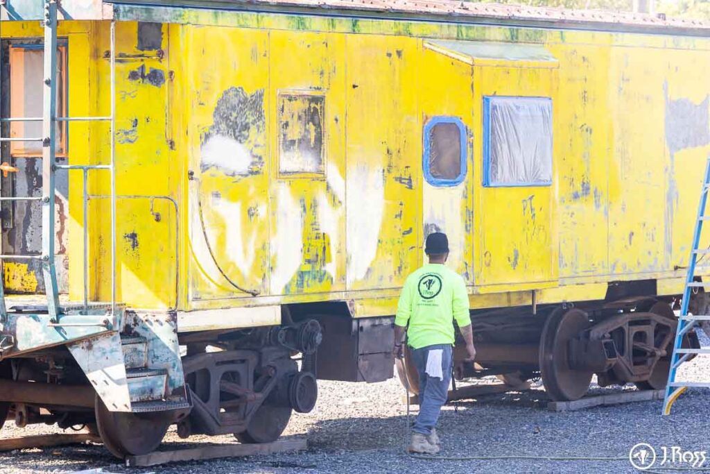 Yellow caboose undergoing paint removal with taped windows during surface preparation by J. Ross crew