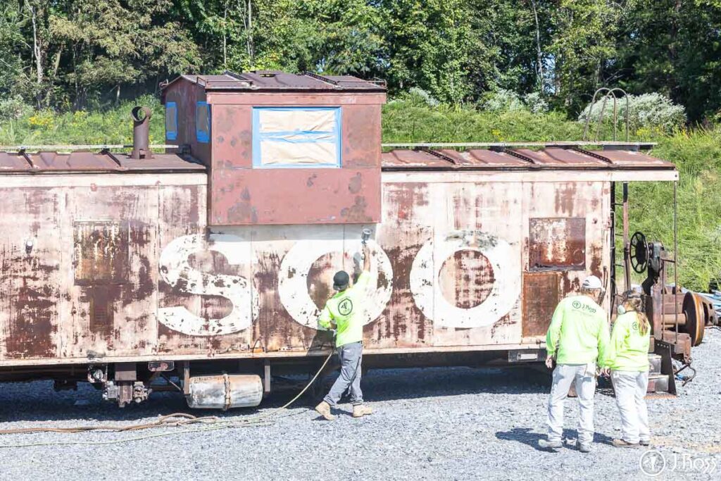 Team applying industrial coatings to rusted steel caboose as part of historic restoration project in Damascus, VA