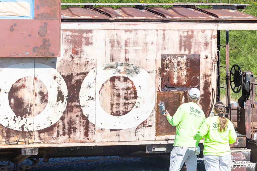 Two crew members in safety gear preparing to spray primer on historic SOO Line caboose exterior