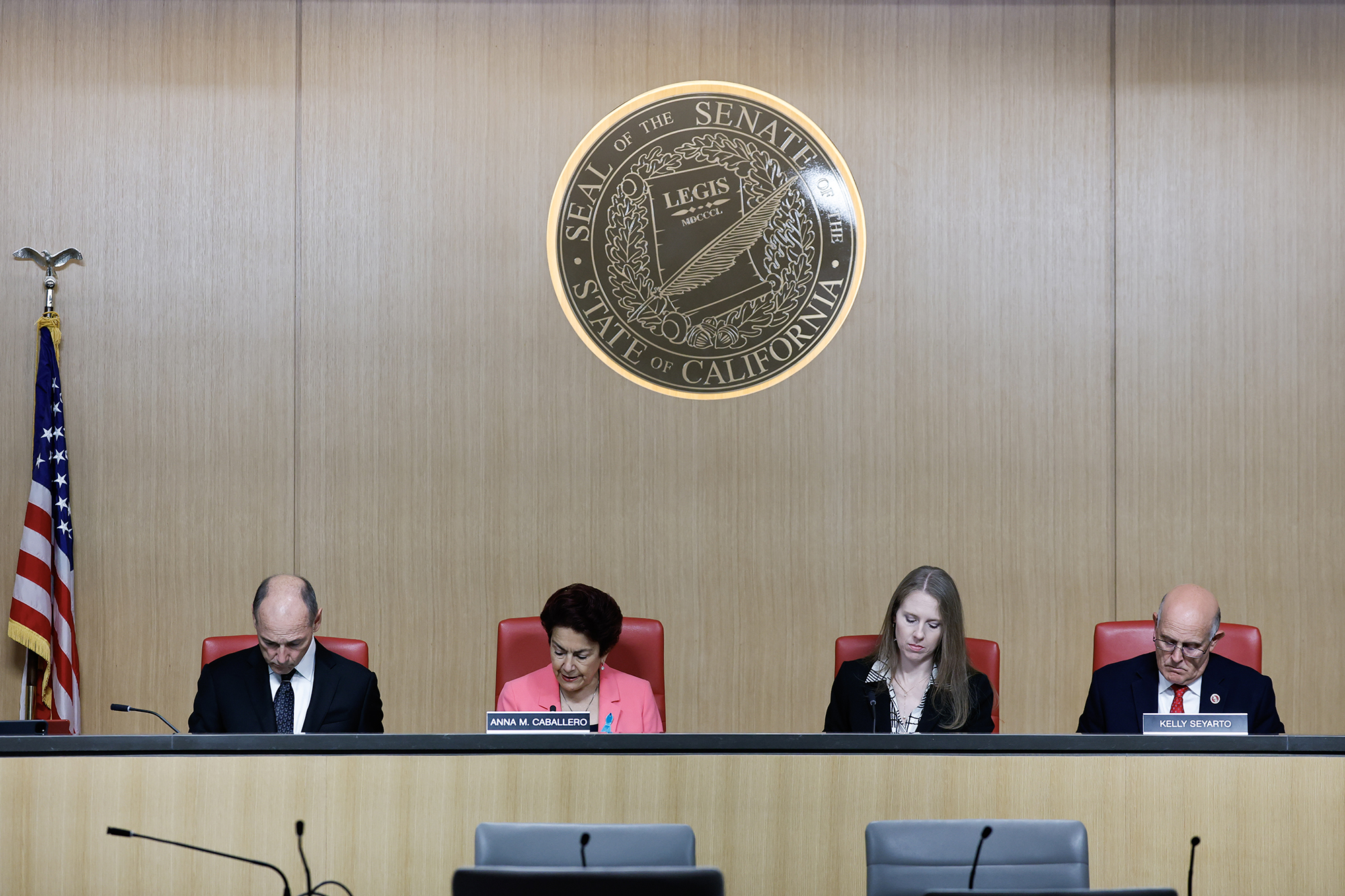 Four lawmakers sit at a long desk under the seal of the California State Senate during a committee hearing. Two men in suits sit on either side, while Senator Anna Caballero, in a bright pink jacket, and a woman in a black blazer sit in the middle. An American flag stands to the left and microphones line the desk in front of them.