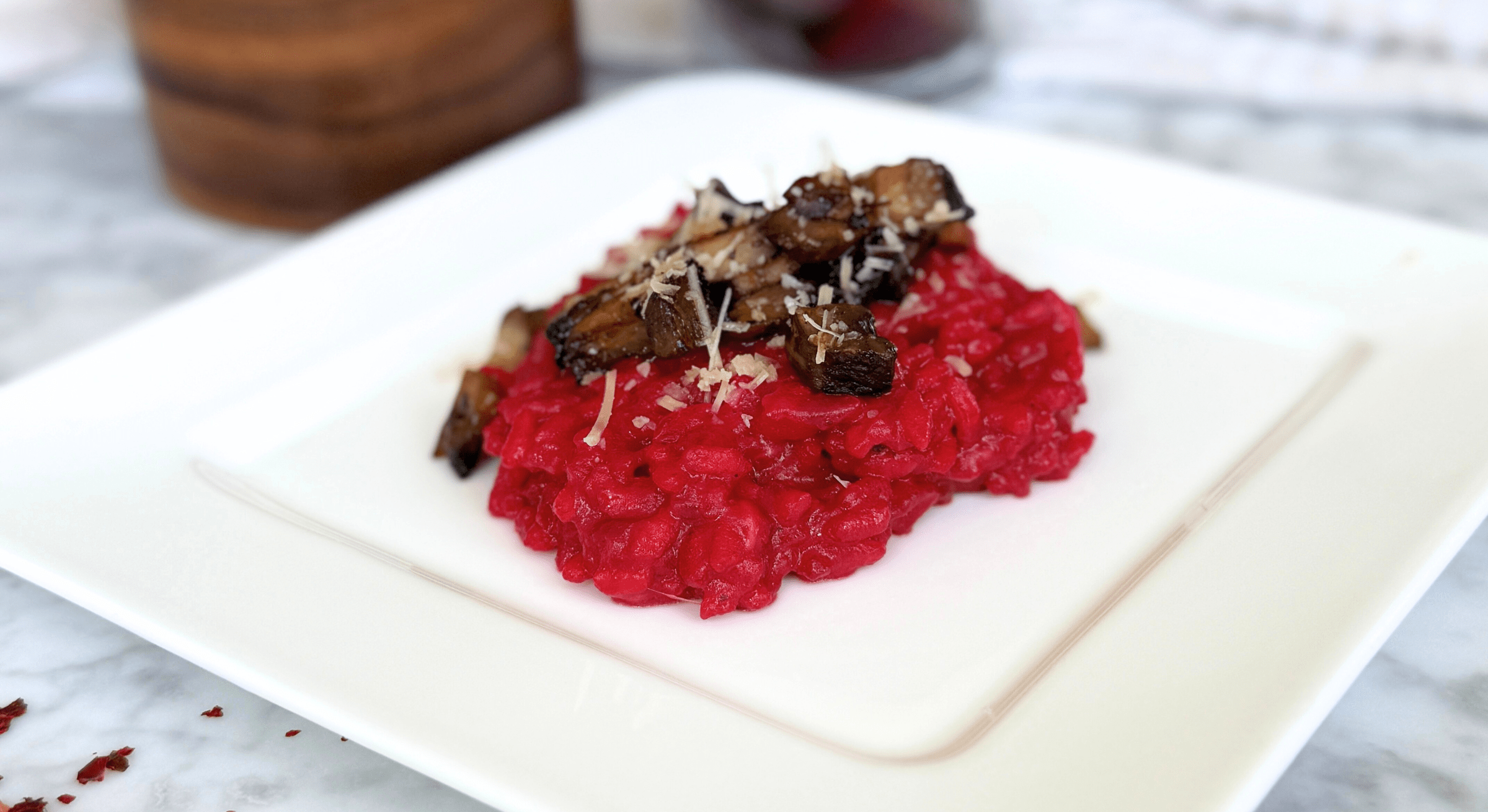 Beet Risotto Recipe. Close-up of bright pink beet risotto topped with mushrooms and cheese, served on a modern square plate.