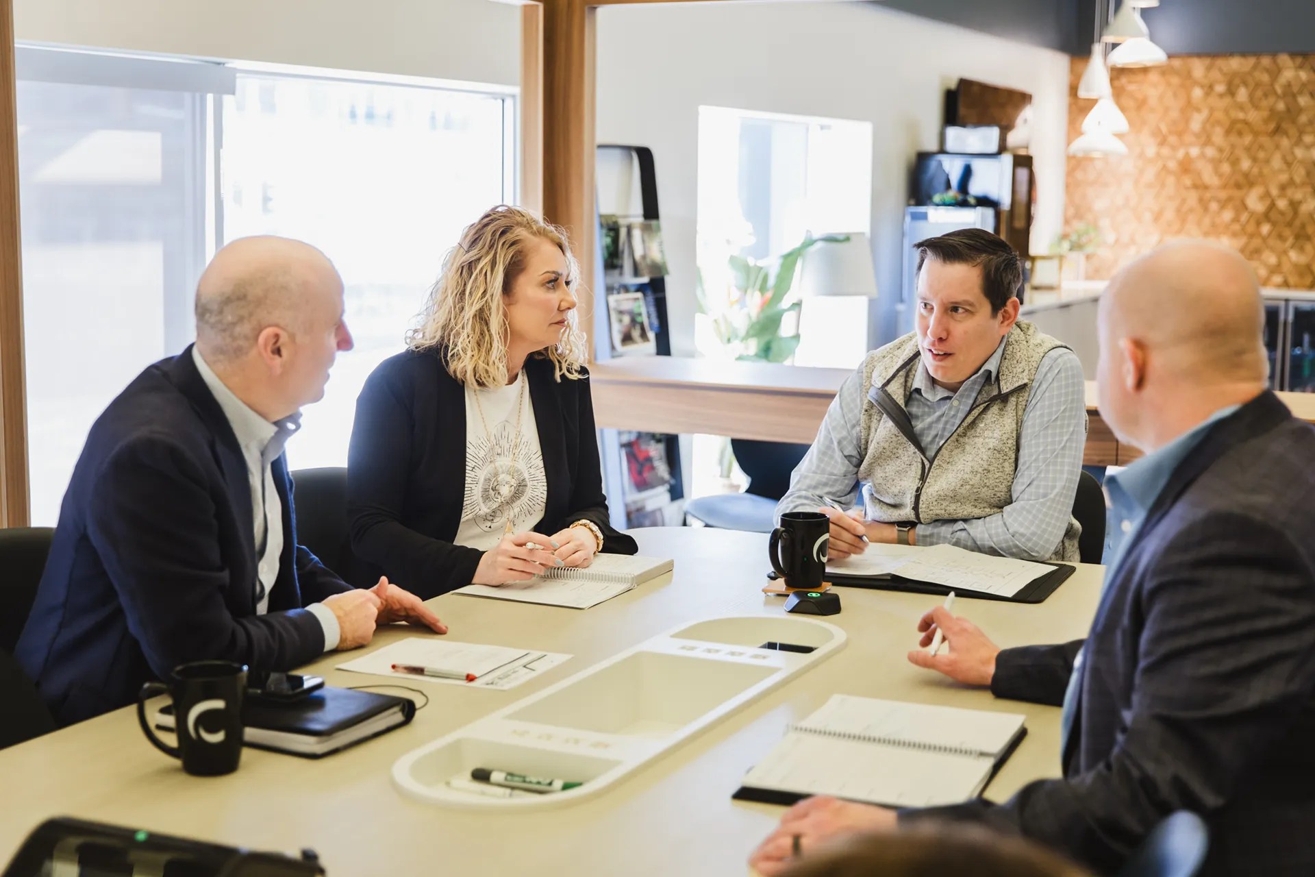 A group of four professionals seated around a conference table, actively discussing ideas and strategies with notebooks and coffee mugs present.
