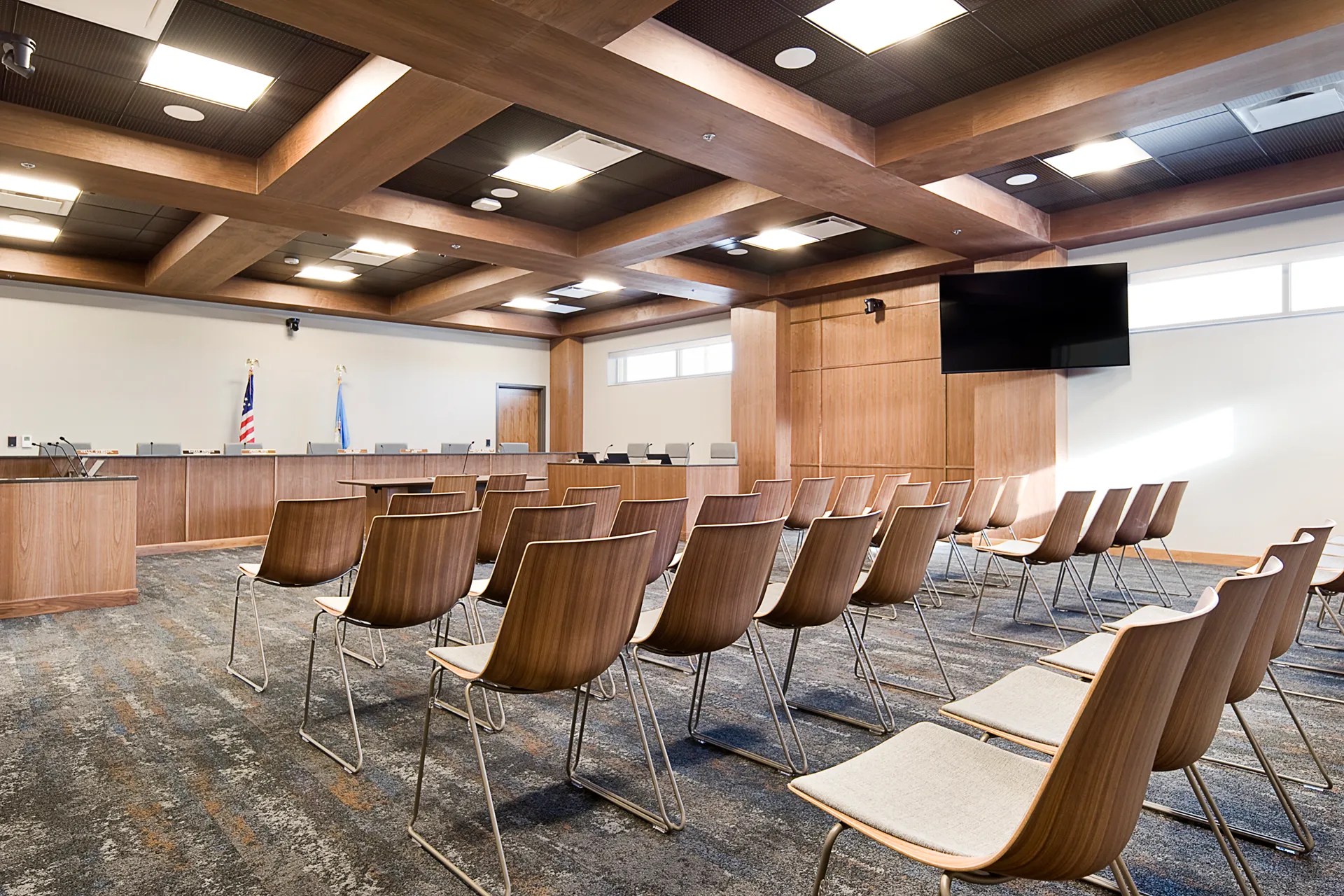 A formal government meeting room with wooden paneling, rows of chairs, and a central table set up for discussions and presentations.