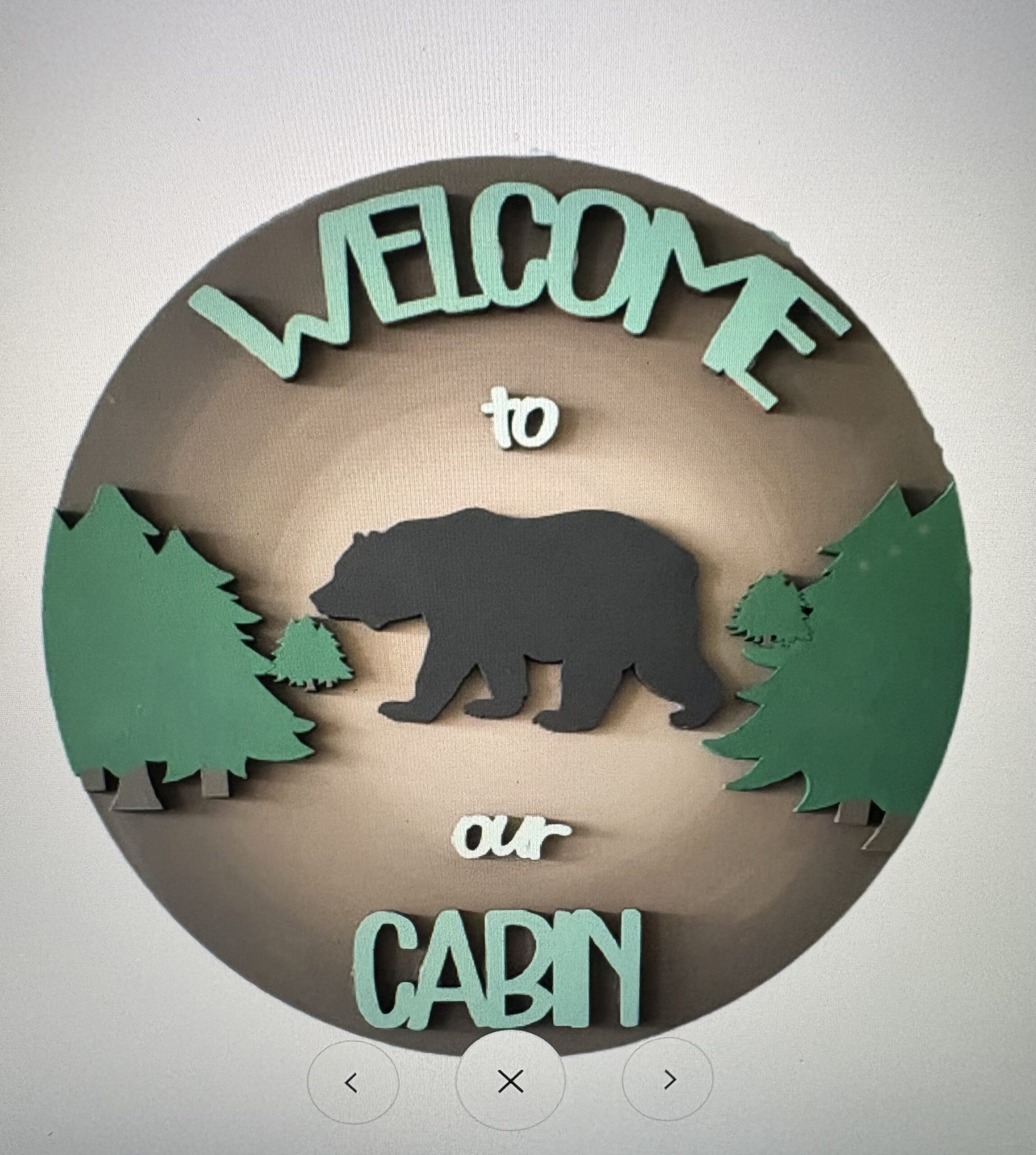 Circular badge with teal 'WELCOME TO' text above a gray bear walking among green evergreen trees on a brown circle background.