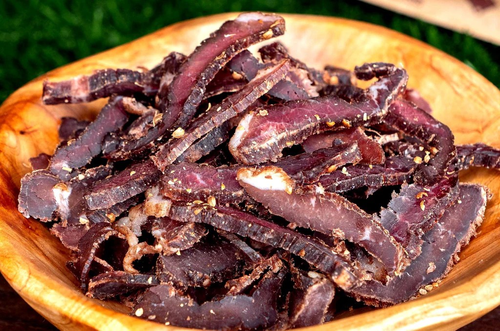 A wooden bowl overflowing with thin slices of traditional South African beef biltong on a green background.