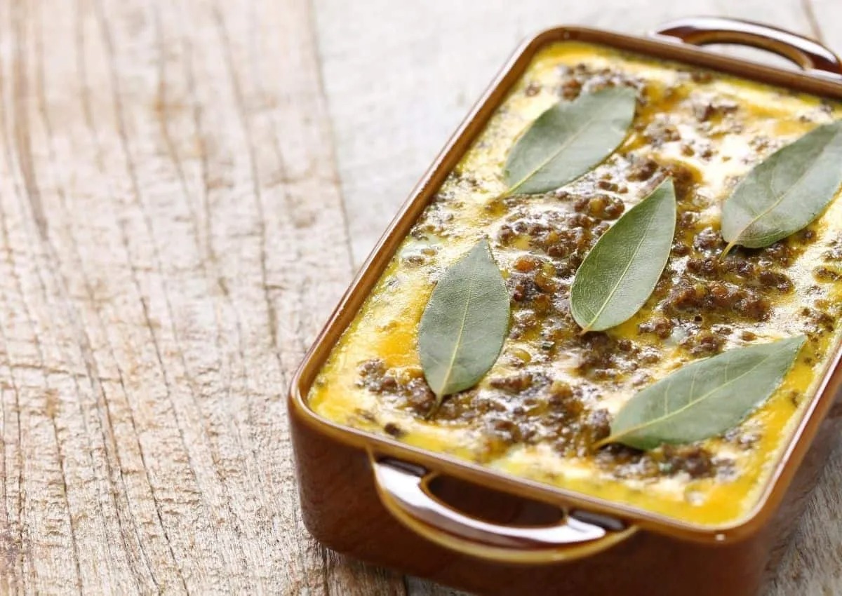 A golden-brown South African bobotie in a white ceramic baking dish, topped with traditional bay leaves and served on an orange cloth.