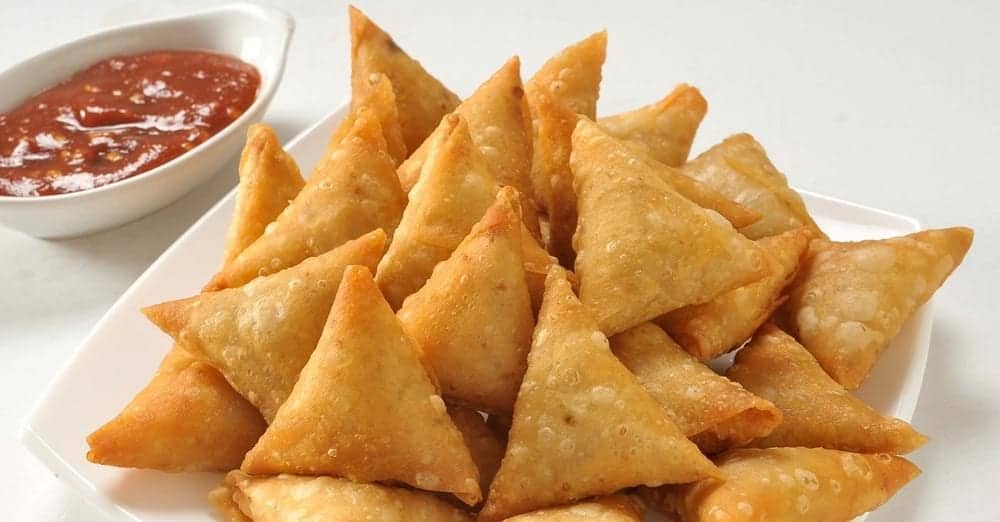 A platter of golden-brown, triangular fried samoosas served with a side of red chili dipping sauce.