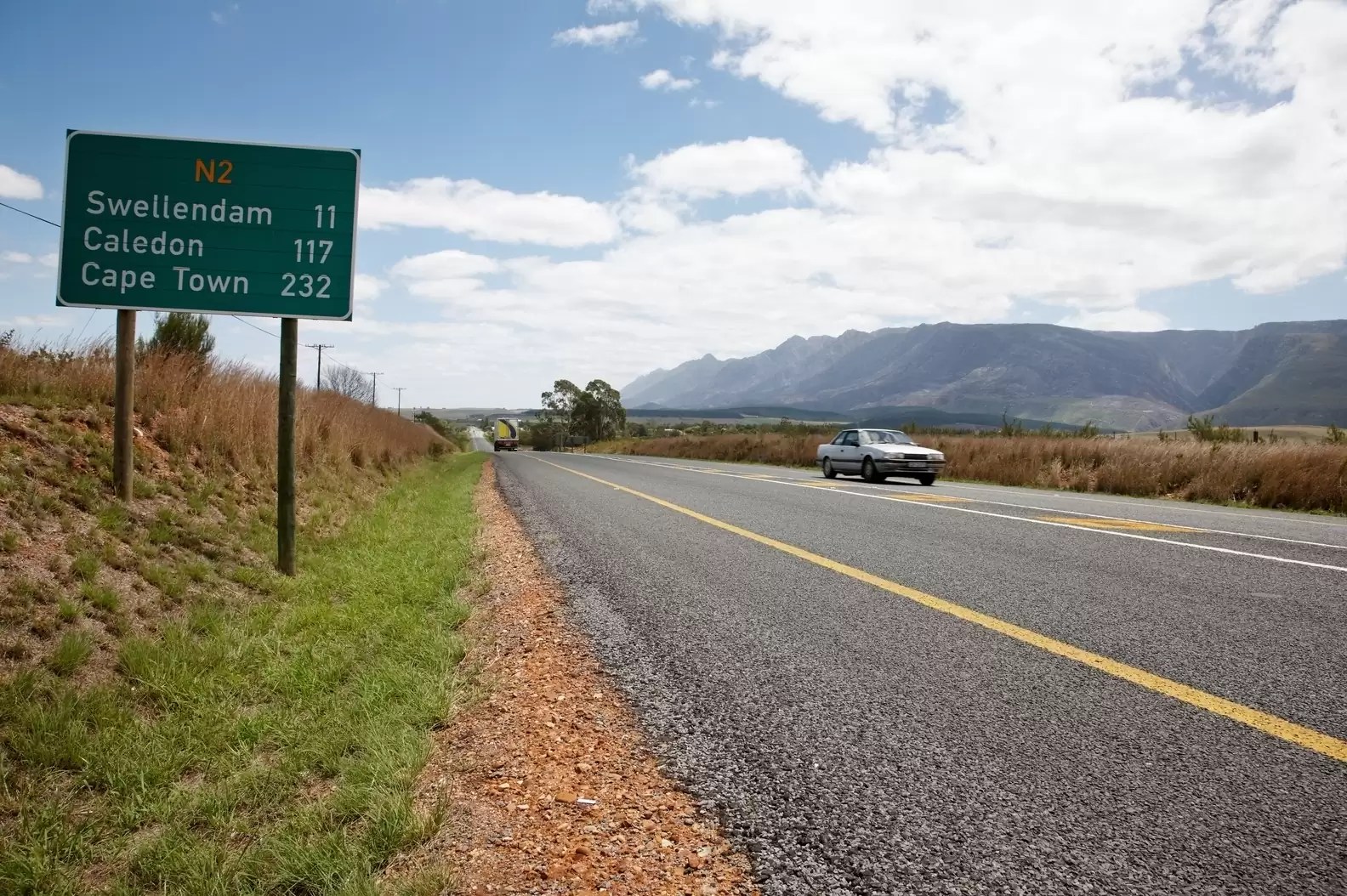 A green highway sign on the N2 in South Africa showing distances to Swellendam (11 km), Caledon (117 km), and Cape Town (232 km) with an open road and mountains in the background.