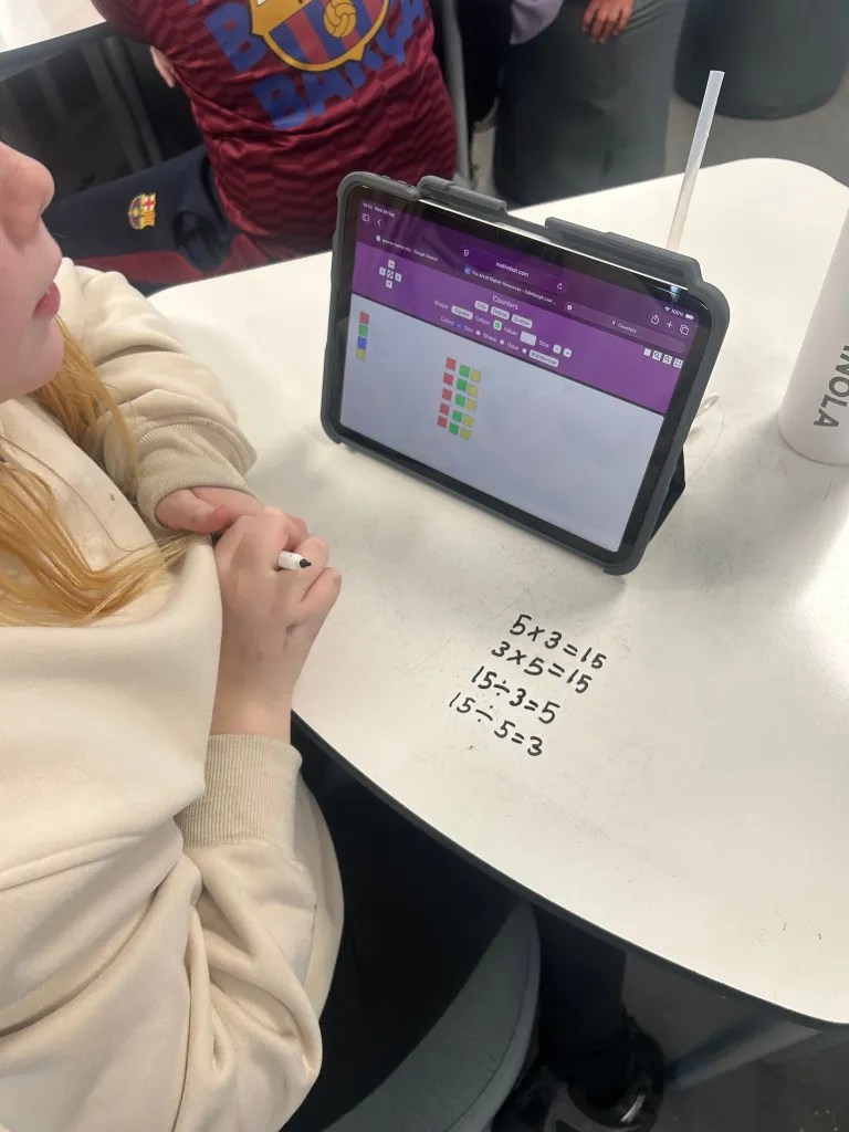A child in a beige sweatshirt using a tablet to solve math problems, with equations written on the table.
