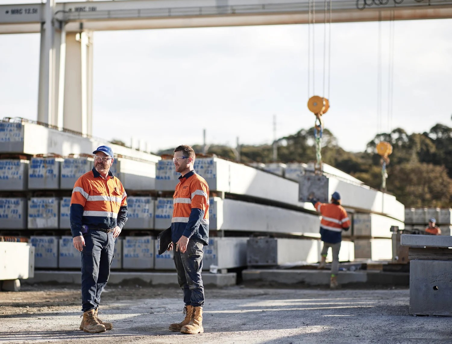 Two construction workers in hi-vis discussing work on a large infrastructure site with concrete beams and a crane in the background.