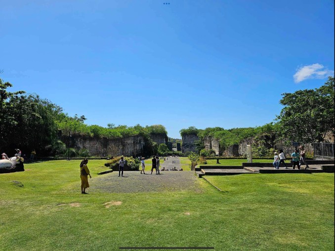 Green grass and open space with large limestone blocks in the background that have greenery growing on top of them at GWK Park Bali