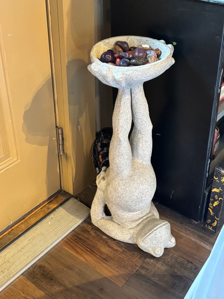 Whimsical upside-down frog sculpture holding a bowl filled with polished stones.