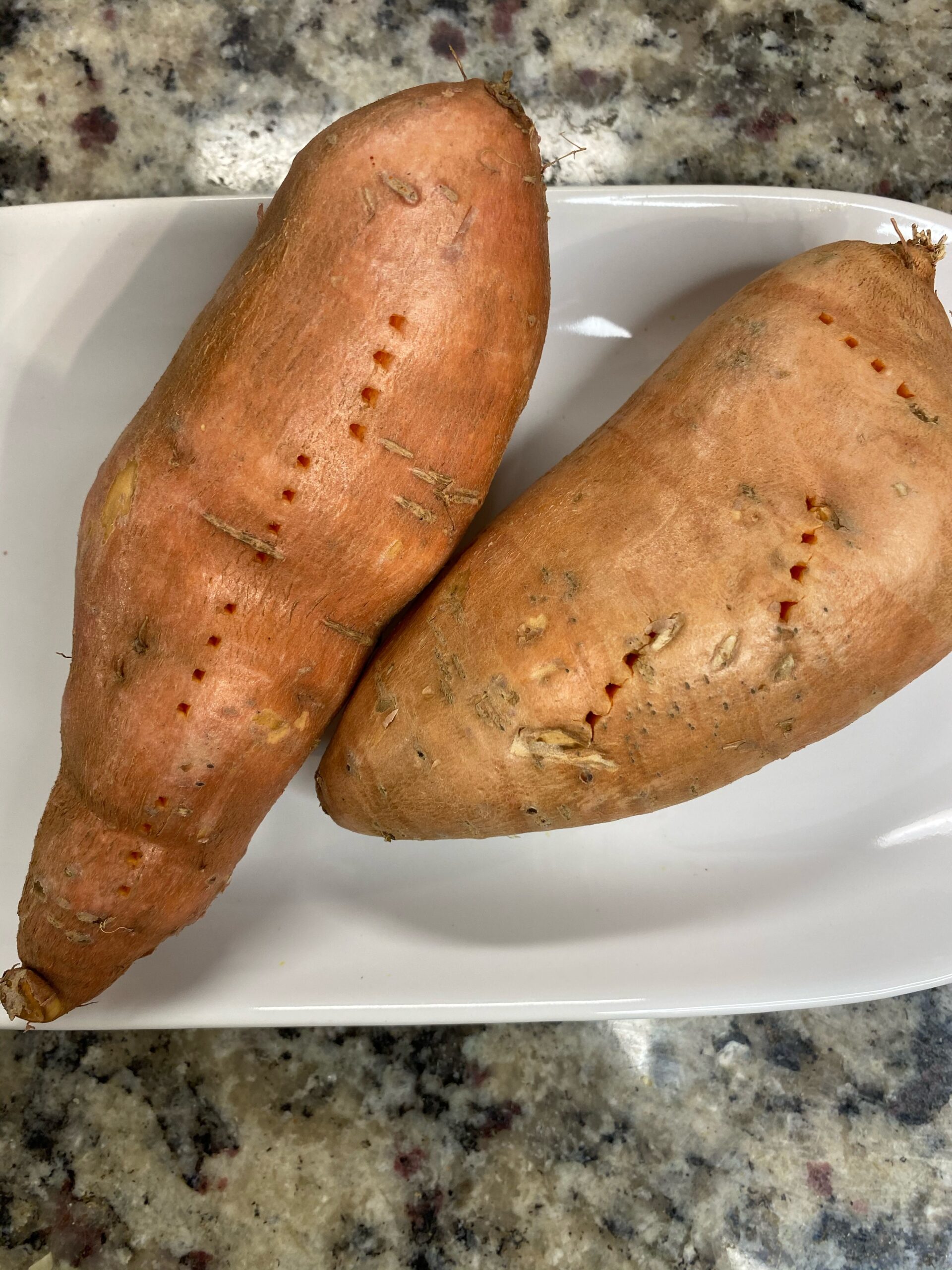 2 sweet potatoes laying on a microwave safe plate. Pierced by a fork and ready to be cooked in the microwave