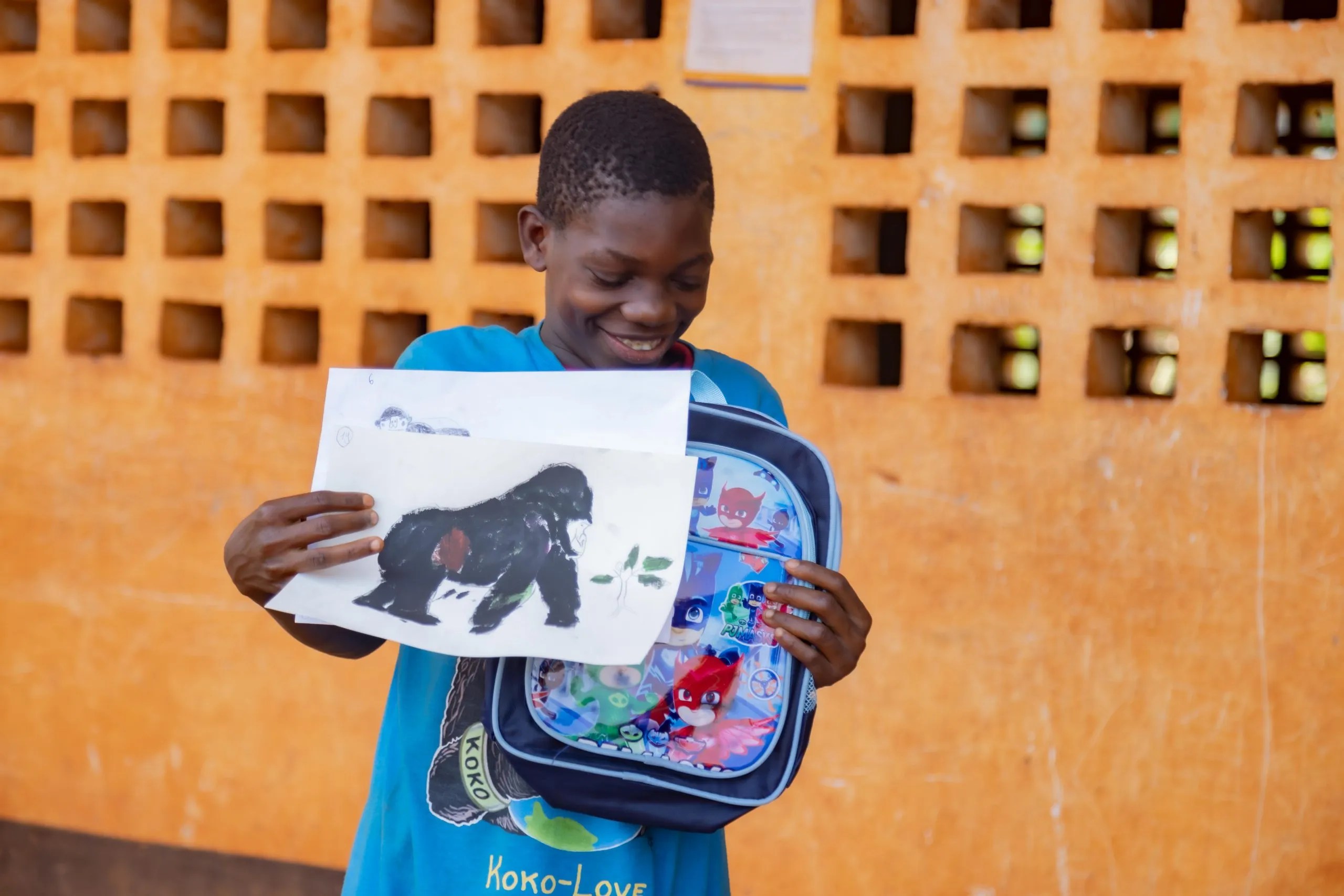 Child smiling holding a backpack with cartoon graphics in one hand and a drawing of a gorilla in the other hand.