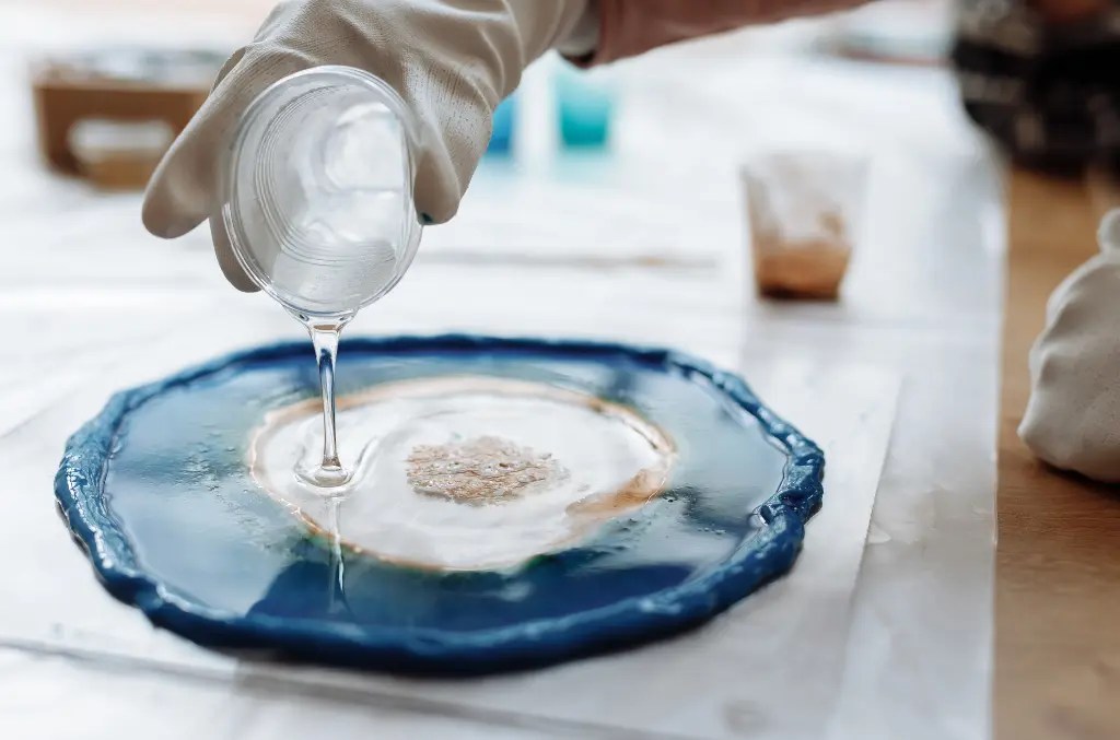 Osoba w rękawiczkach wylewa płynną żywicę epoksydową z kubeczka na okrągłą formę w kolorze niebieskim, tworząc dekorację DIY w technice rękodzieła.