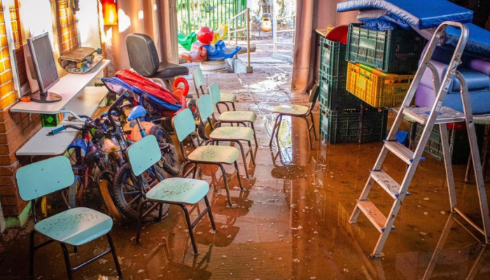 Interior de uma sala de aula de educação infantil alagada, com o chão coberto por água barrenta. Cadeiras escolares, brinquedos e caixotes estão empilhados e amontoados, evidenciando os danos causados por enchente em uma escola em área de risco.