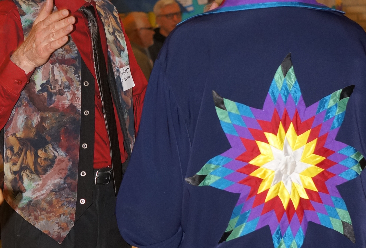 White man in colourful vest and Indigenous man in colourful ribbon shirt having a conversation