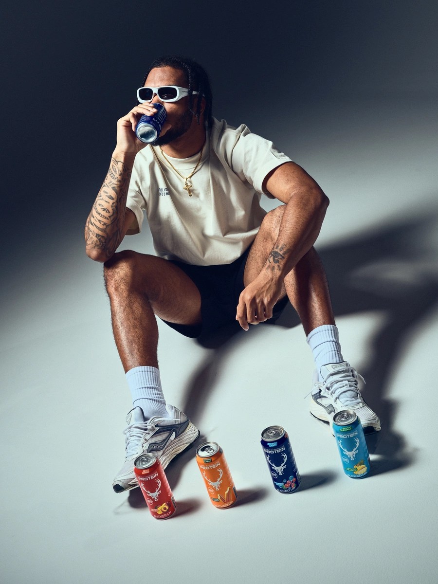 A young man with dark skin, tattoos on both arms, and dreadlocks is squatting on a plain, light-colored studio floor against a dark gradient background. He is wearing white sunglasses, a loose beige T-shirt with small text on the chest, black shorts, white athletic shoes, and white socks. He is holding a blue soda can to his mouth with his right hand, and his left hand rests on his knee. Around his feet are four colorful cans of protein drinks in red, orange, blue, and teal, each with a deer logo and the word 'PROTEIN' on them. The lighting is soft and directional, creating subtle shadows and highlights that emphasize his relaxed pose and casual style, evoking a modern, lifestyle photography vibe.