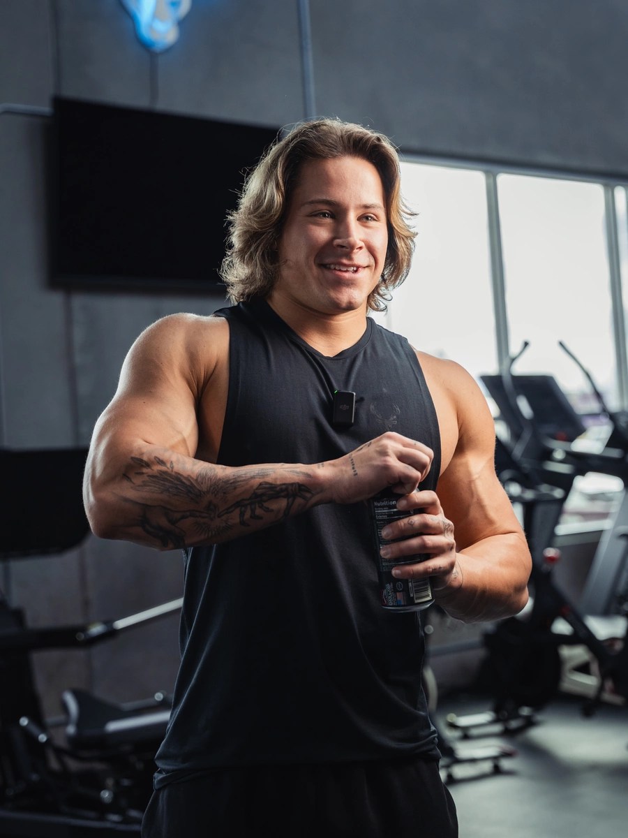 A young man with shoulder-length wavy blonde hair, wearing a sleeveless black athletic shirt, is smiling while holding a protein bar in a gym. His muscular arms are visible, with a detailed tattoo of a tree and branches on his left arm. The gym background features large windows, exercise equipment, and a dark wall with a mounted flat-screen TV. The lighting is bright and natural, highlighting his cheerful expression and athletic physique, with a professional lifestyle photography style.