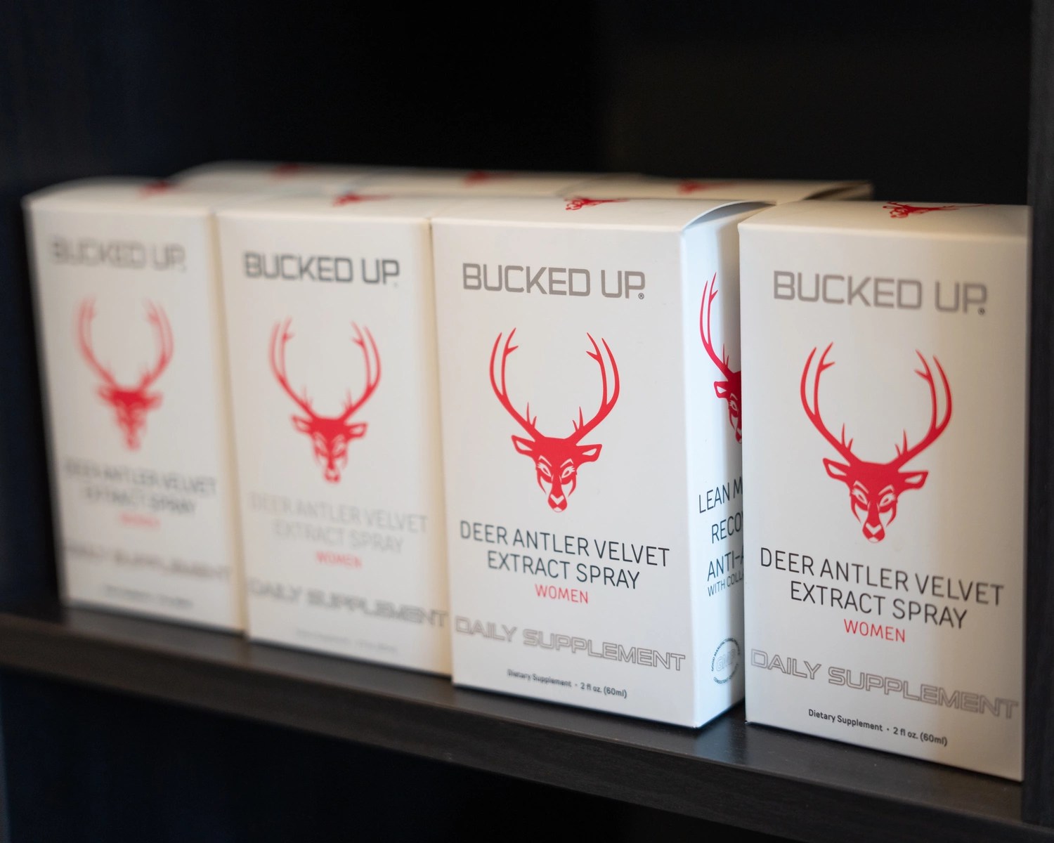 A row of white boxes of Bucked Up Deer Antler Velvet Extract Spray for women, featuring a red deer head logo with large antlers, arranged neatly on a black shelf. The boxes are aligned side by side, with the front box slightly more prominent, showing the product name and details. The background is dark, emphasizing the clean, minimalist design of the packaging, with soft lighting highlighting the boxes' surfaces.
