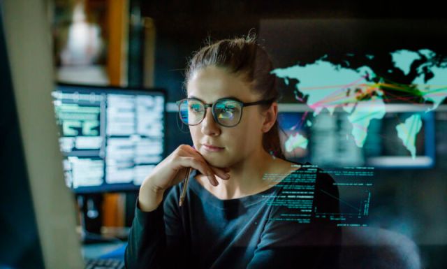 Stock Image - Young woman wearing glasses surrounded by computer monitor in a dark office.  In front of it is a transparent display showing a map of the world with some data.