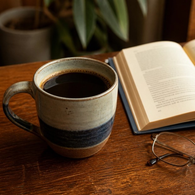 A ceramic mug of dark roasted coffee on a wooden table beside an open hardcover book and reading glasses