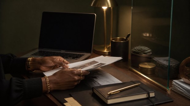 Black woman’s hands reviewing printed pages beside a laptop with a blank screen, brass pen, notebook, and warm desk lamp