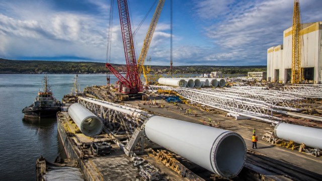 A high-resolution editorial photograph of Hudson River port and renewable energy manufacturing infrastructure under construction in the Albany region, featuring heavy-lift dock works, steel tower sections, cranes, and workers.