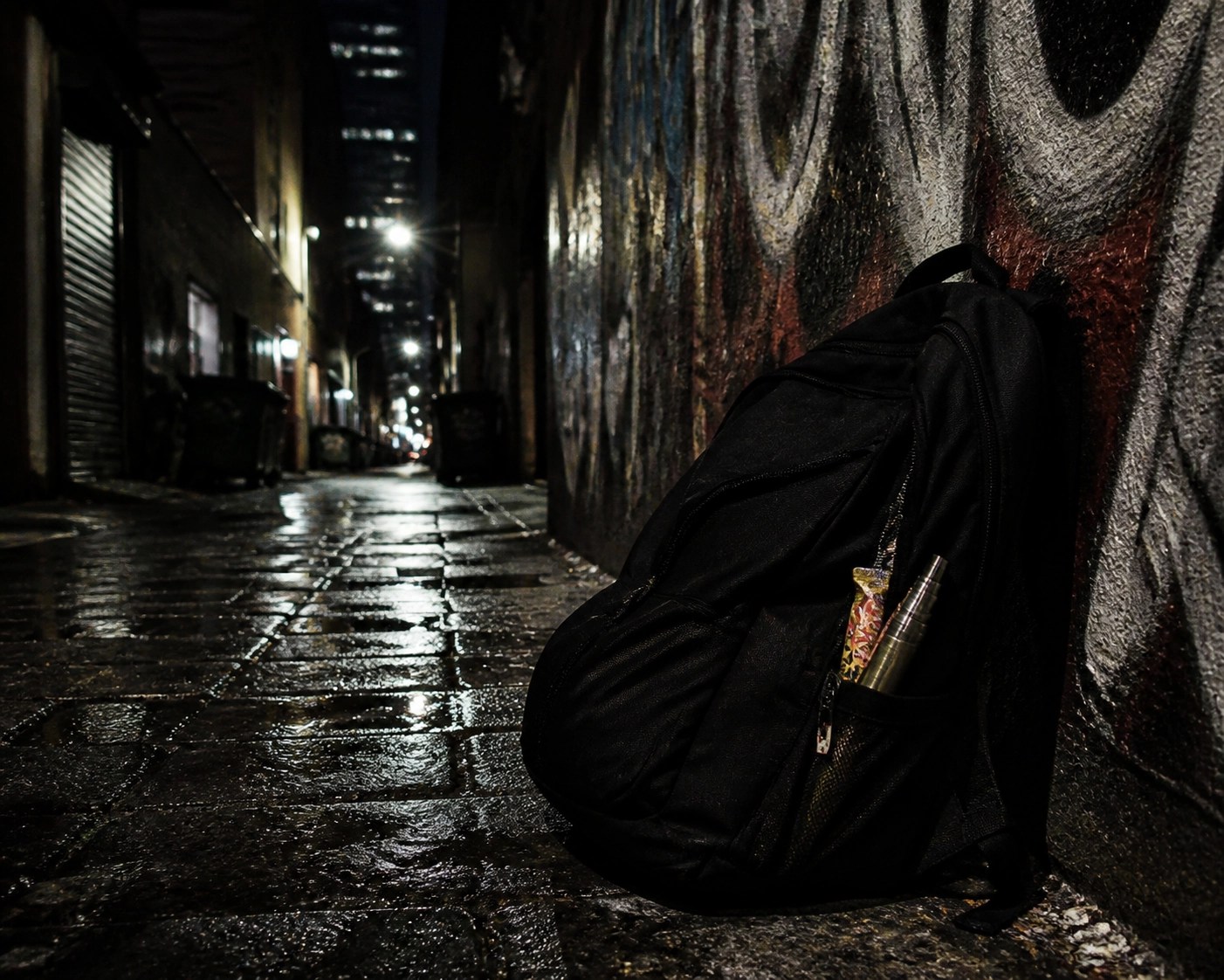 A black backpack in a gritty NYC alley containing a hidden vape pen and fake candy wrappers.