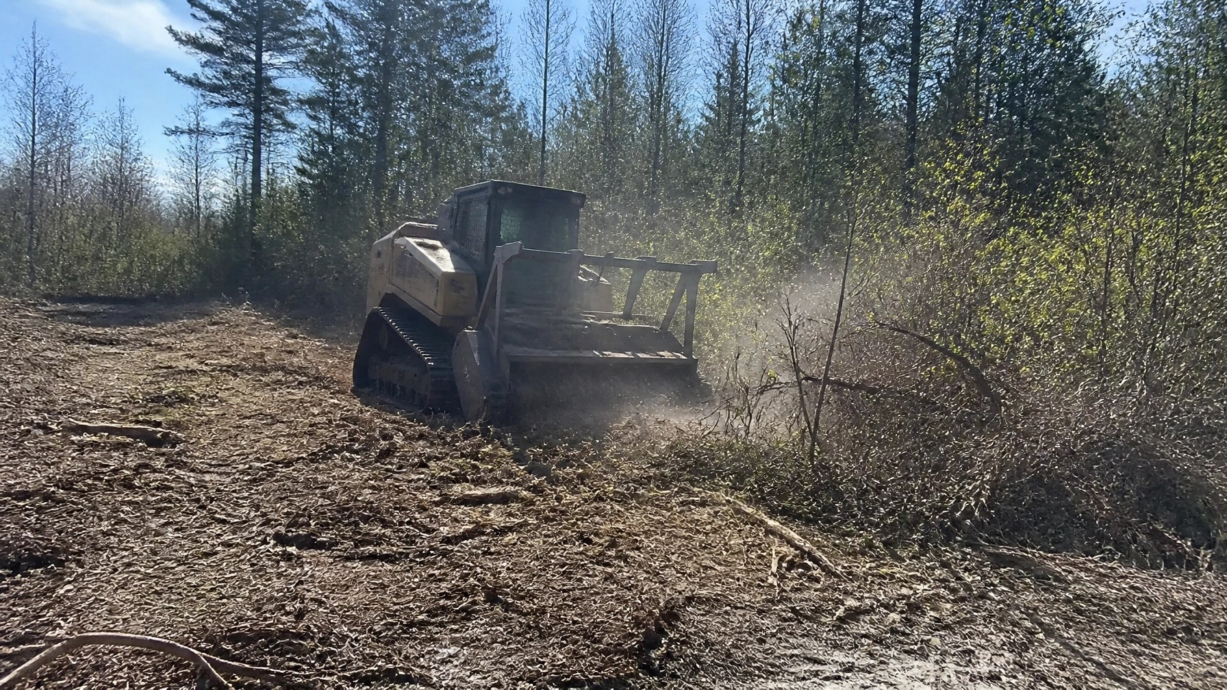 Authentic field photograph of brand-neutral forestry mulching equipment working on a commercial site in daylight. Captured from ground level with a real-world handheld feel, showing mulch, mud, dirt, and raw site conditions. Slightly imperfect composition reinforces documentary-style field realism.