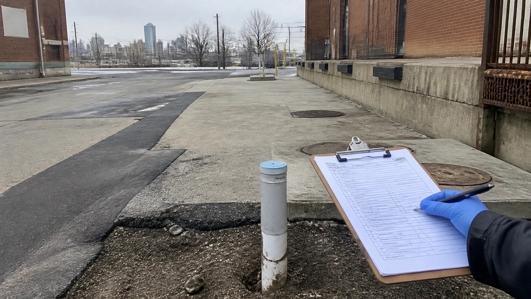 Field reconnaissance scene with a capped monitoring well, industrial loading area, site checklist, and visual indicators of former fuel and fill conditions