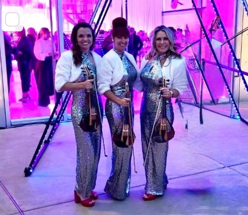 Three members of Urban Electra in matching silver sequin jumpsuits, holding electric violins in front of a geometric stage setup illuminated by vibrant purple event lighting.
