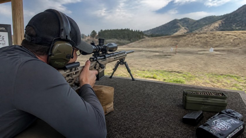 A shooter at a rifle station aiming downrange with a scoped rifle, with targets and range gear visible